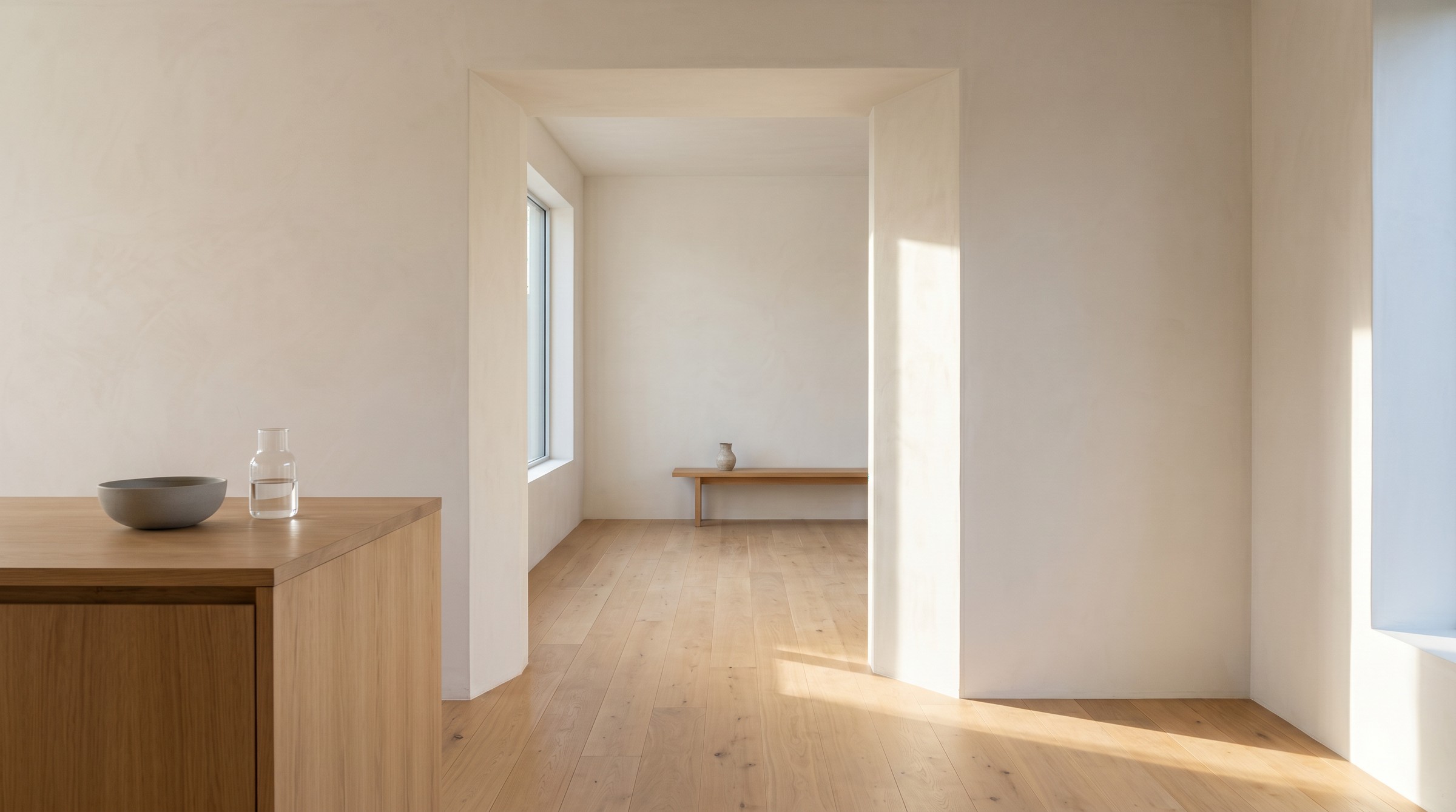 A quiet domestic threshold — kitchen island corner, deep wall reveal between two oak-floored rooms, morning light raking across the floor.